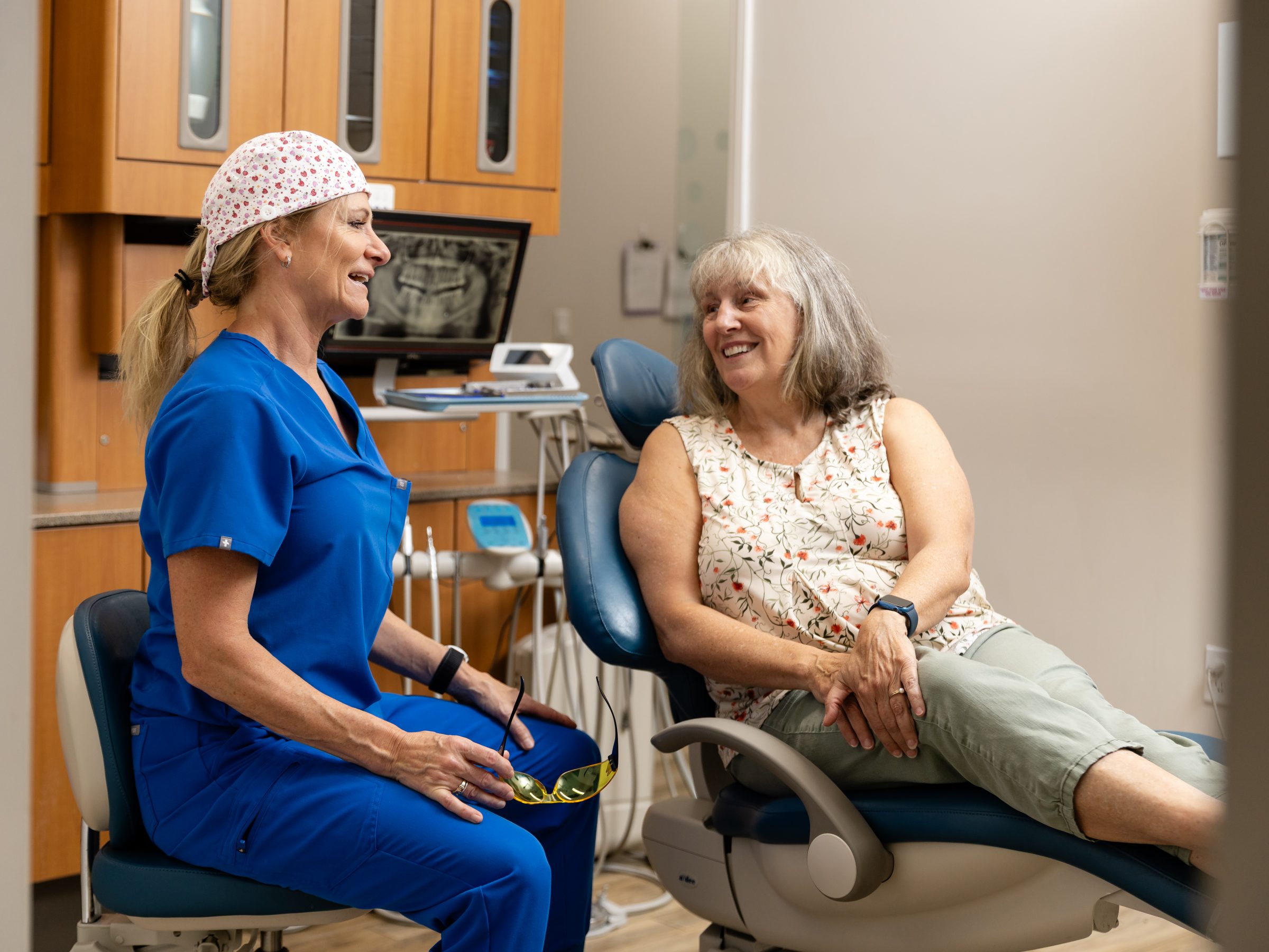 A person in blue scrubs and a seated person converse happily in a modern dental office, with dental equipment in the background.
