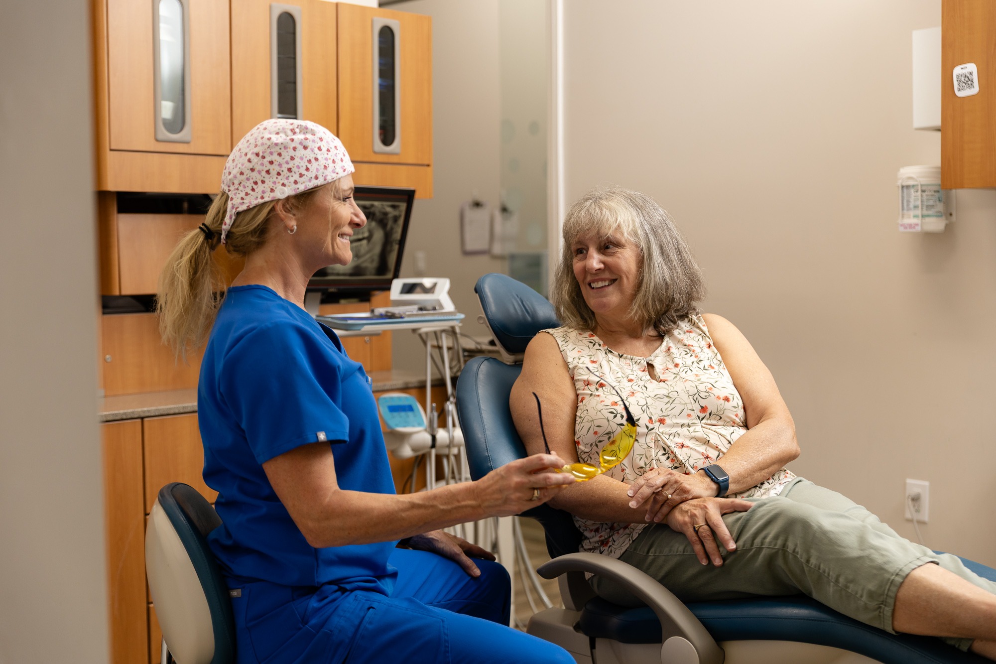 Two people in a dental office, one in scrubs and the other seated in the exam chair, smiling at each other.