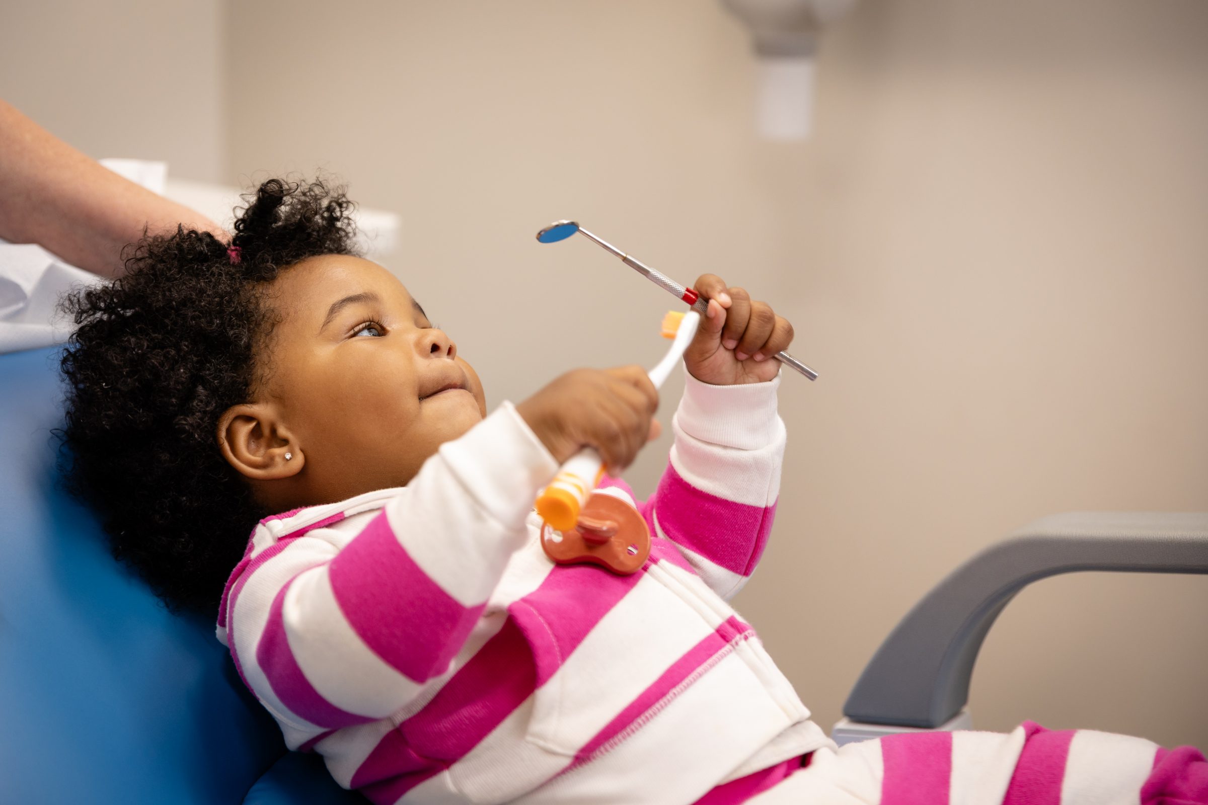 A toddler sits on a chair holding a toothbrush and dental tool, wearing a pink and white striped outfit. A person assists.