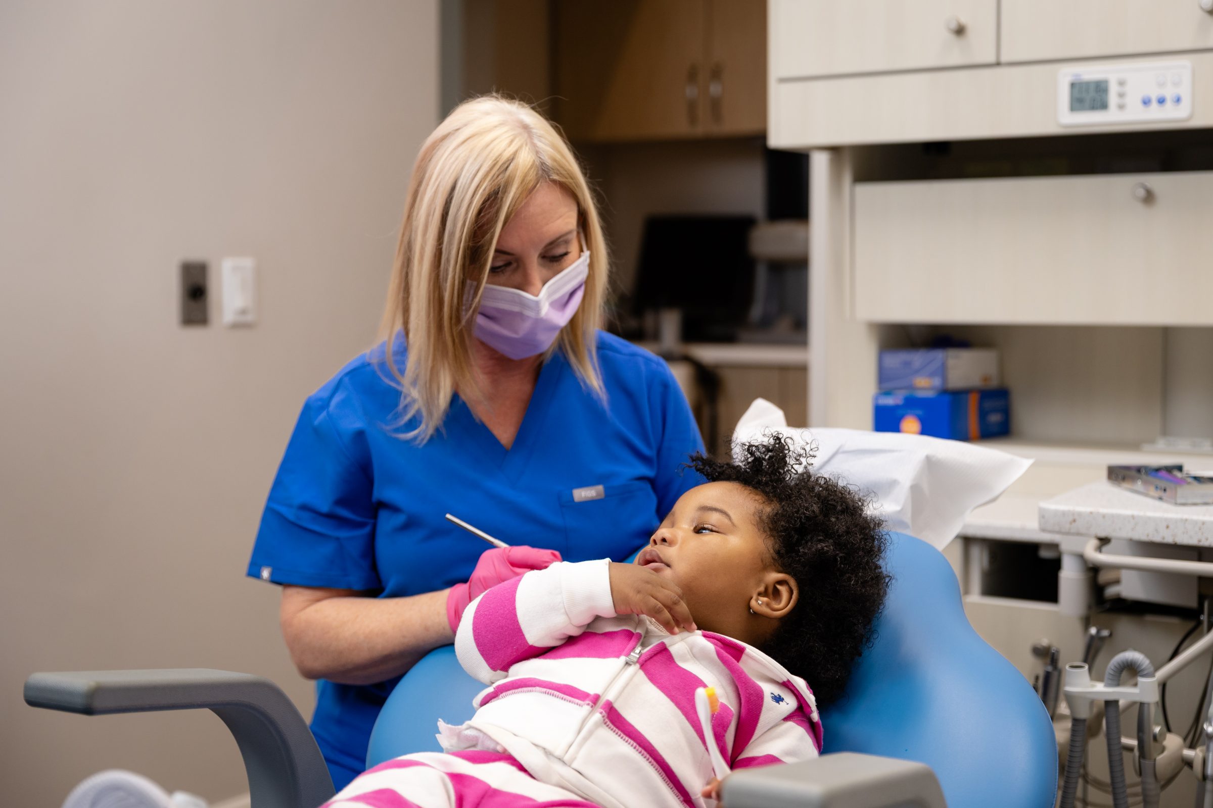 A person in blue scrubs attends to a child in a dentist's chair. The child holds a toothbrush, and the environment is clinical.