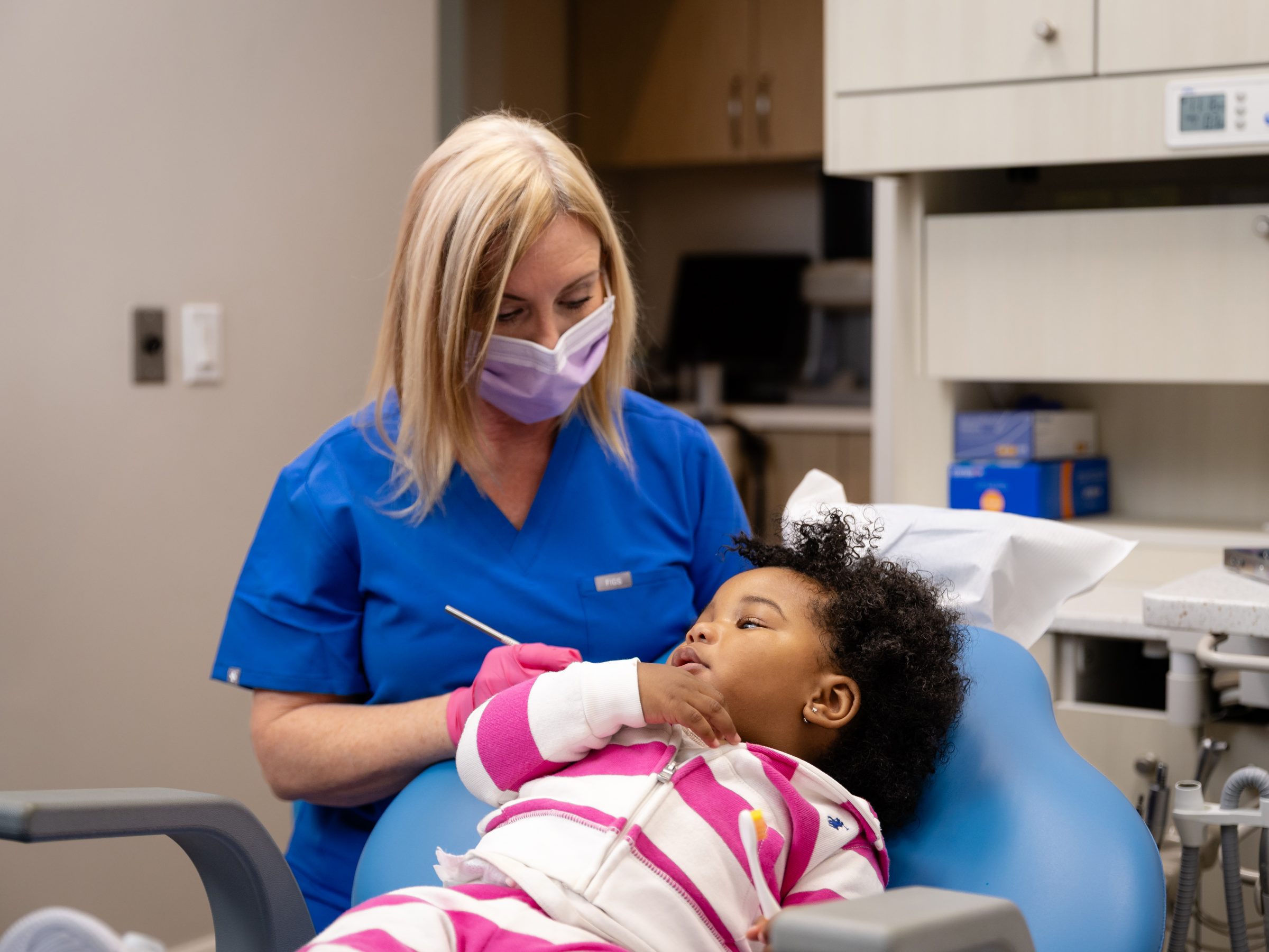A person in blue scrubs assists a child in a dental chair. The setting appears to be a dental clinic.