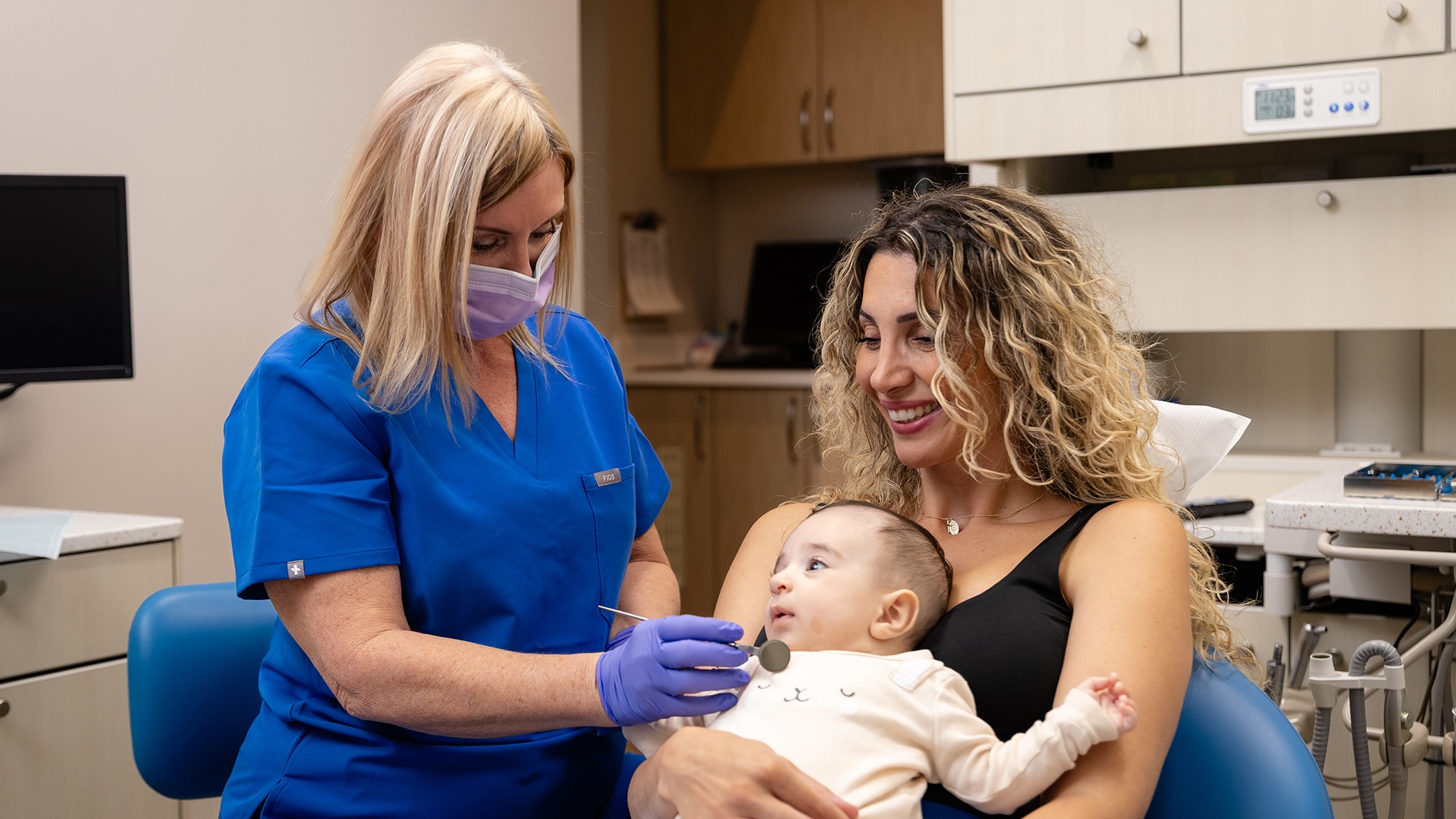 A person in blue scrubs and mask attends to a smiling person holding a baby in a dental office setting.