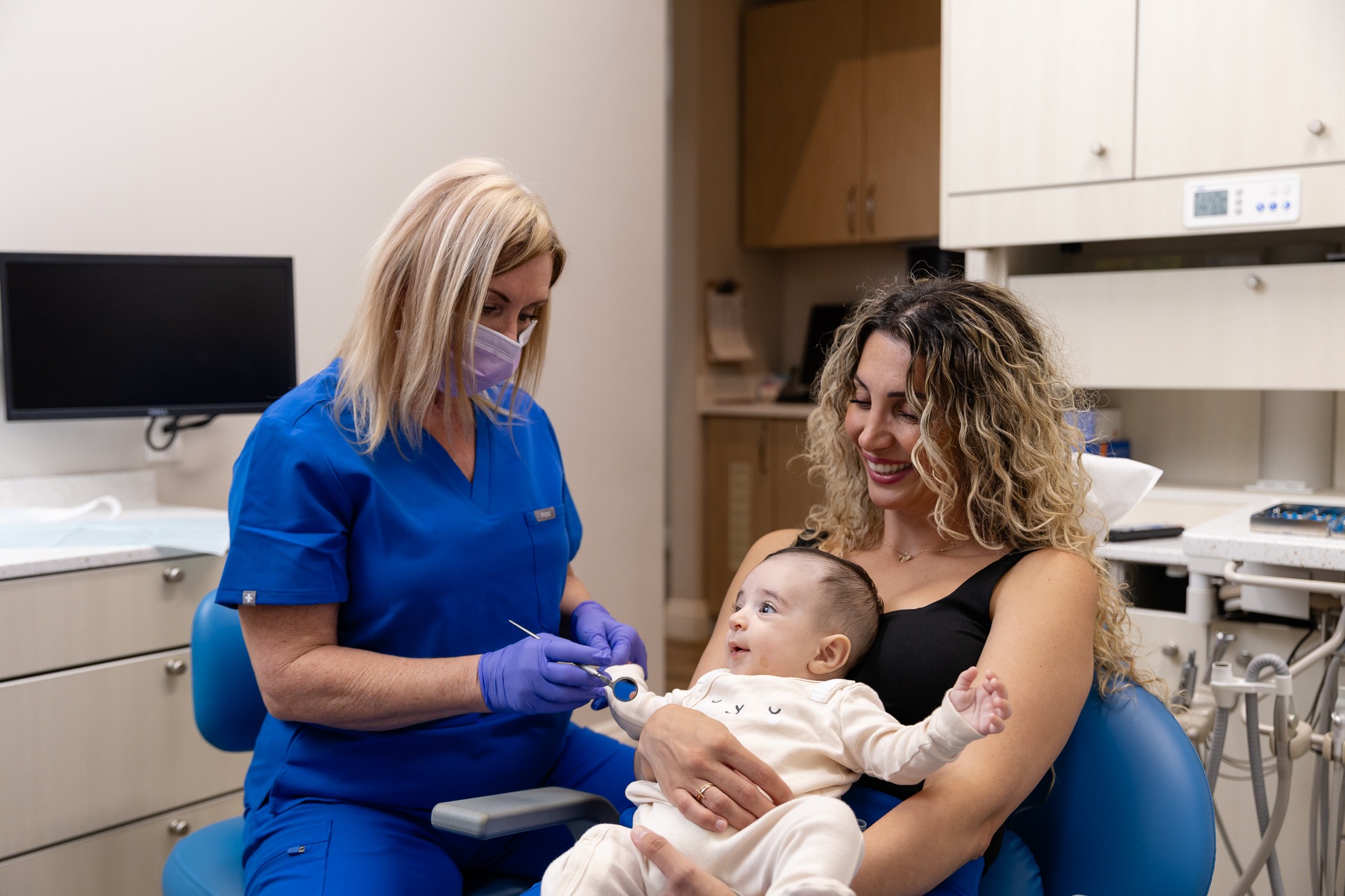 A person in blue scrubs smiles while examining a happy baby sitting on another person's lap in a dental office.