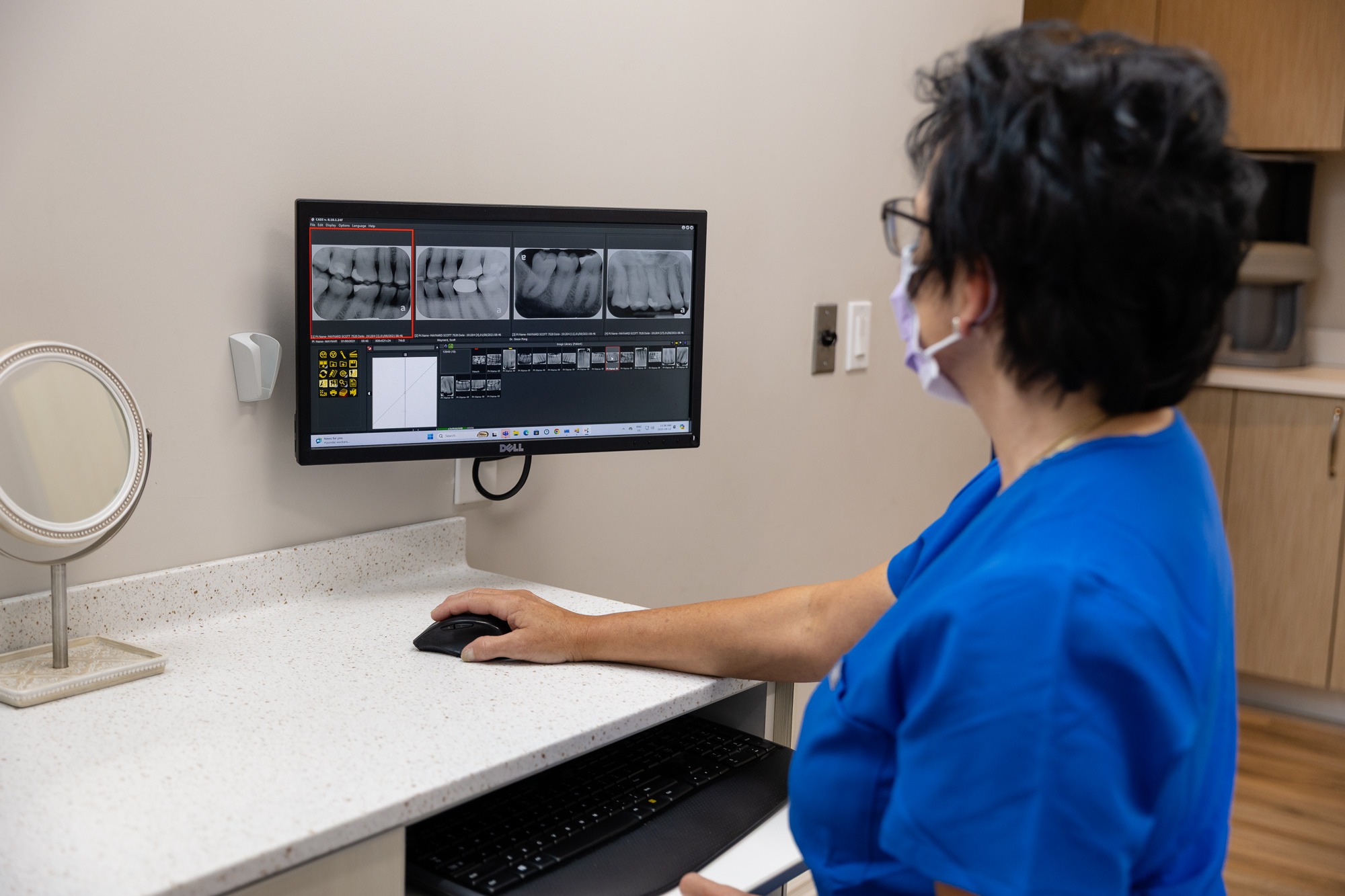 A person in scrubs examines dental X-rays on a computer screen in a clinical setting, hand on mouse, tabletop mirror present.