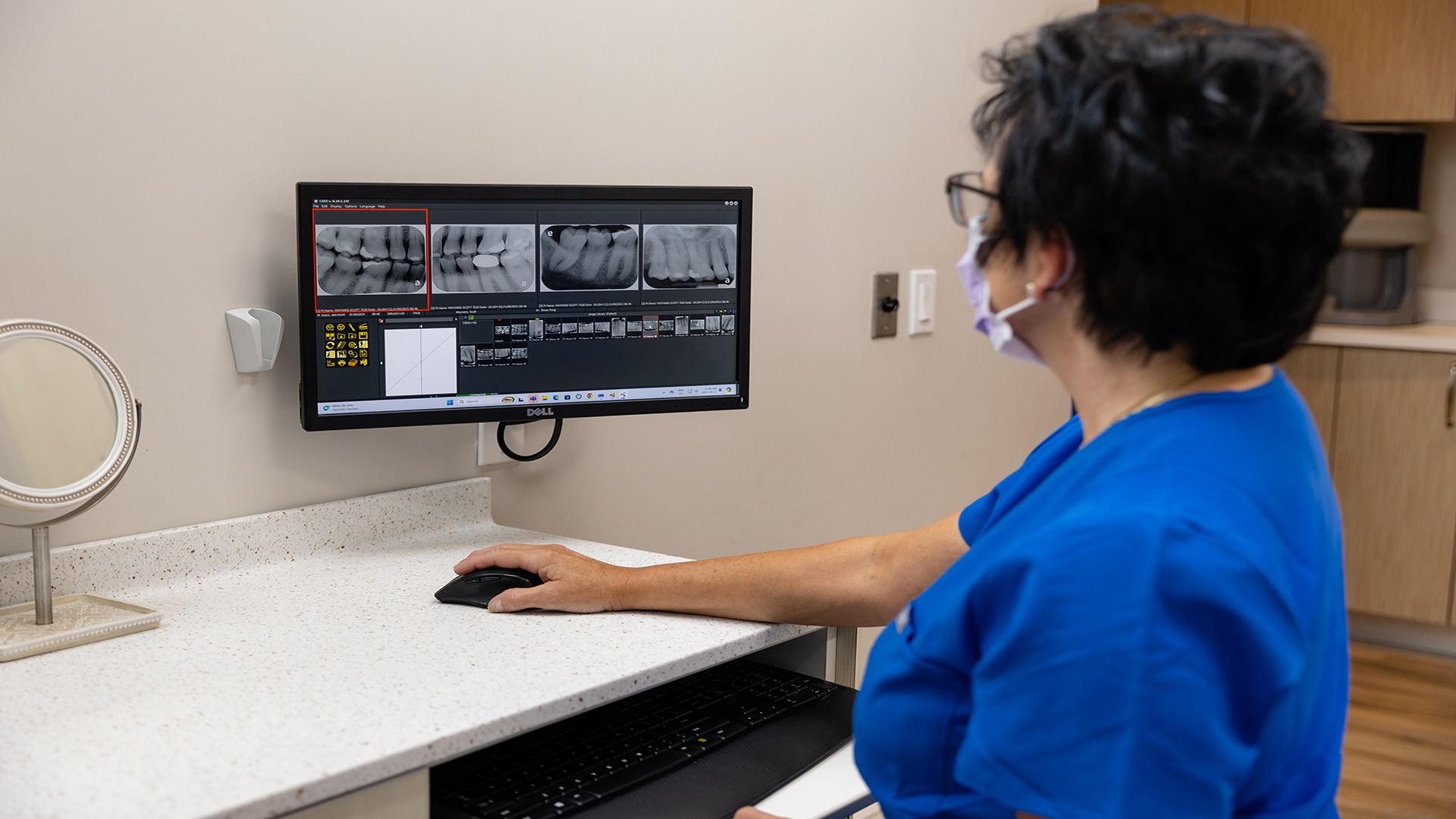A person in blue scrubs examines dental X-rays on a computer screen in a clinical setting, using a mouse for navigation.