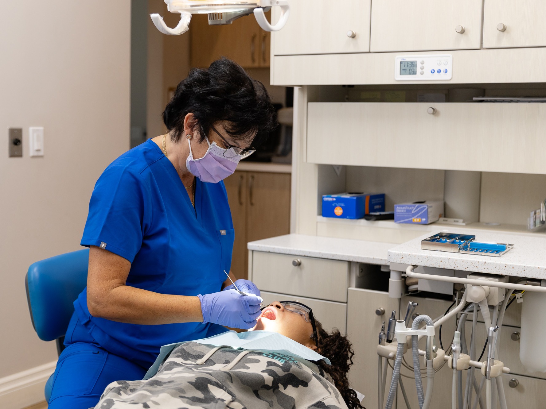 A person in blue scrubs and mask is performing dental work on another person in a modern dental office equipped with various tools.