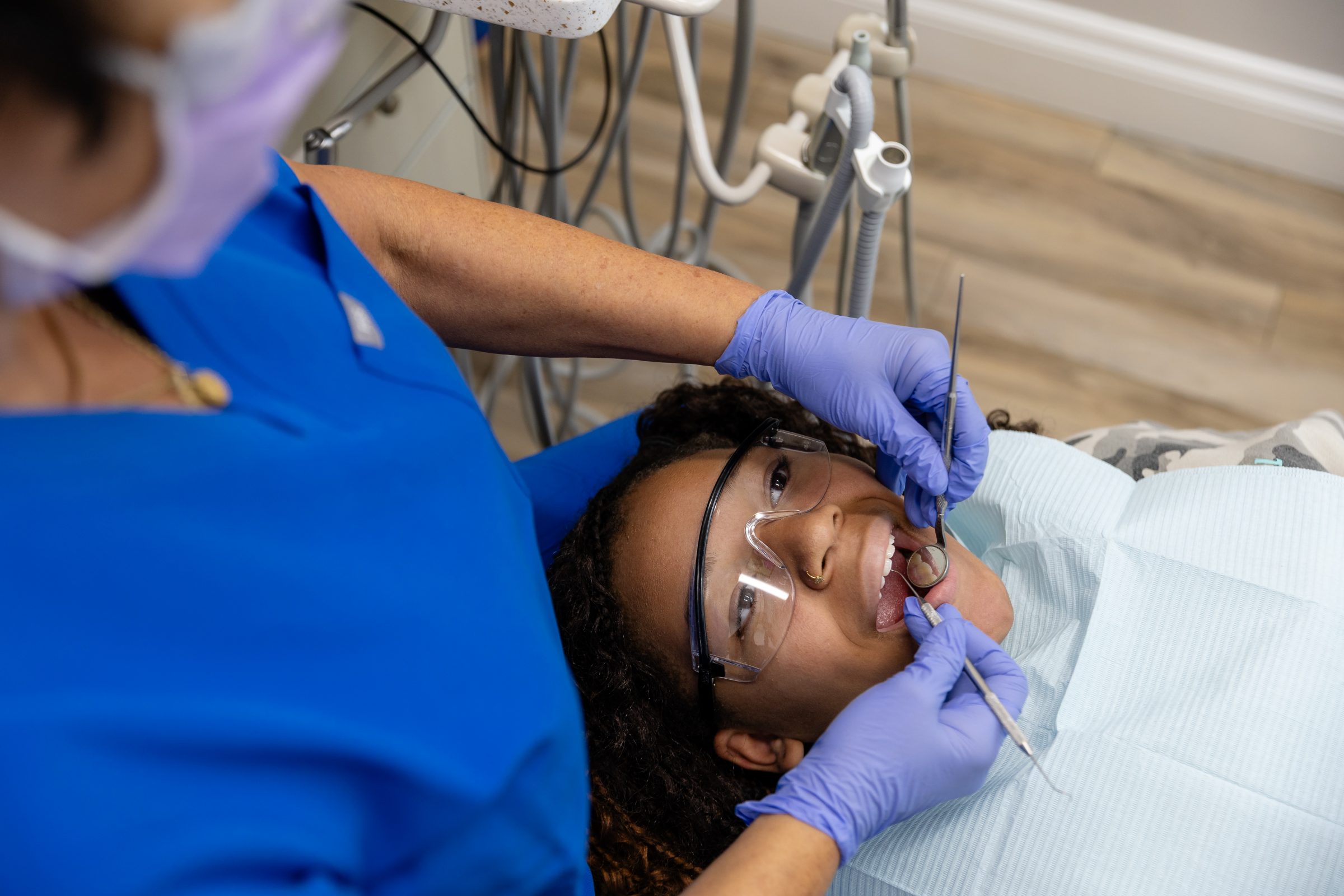 A person in a dentist's chair receives a dental check-up, wearing protective glasses and a bib. Dental tools and gloves are visible.