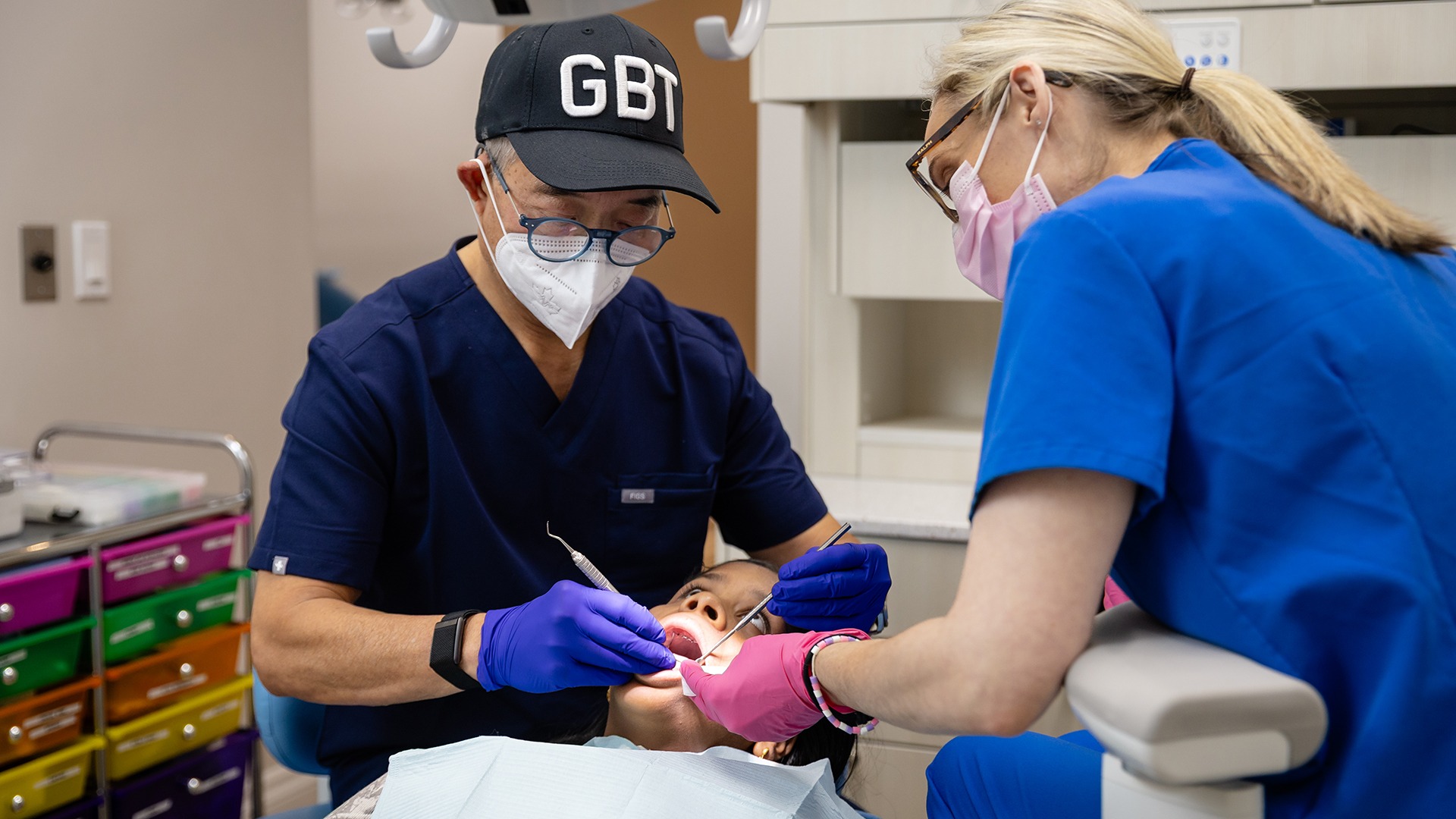 Two dental professionals perform a procedure on a patient, using tools. Colorful storage drawers are visible in the modern dental clinic.