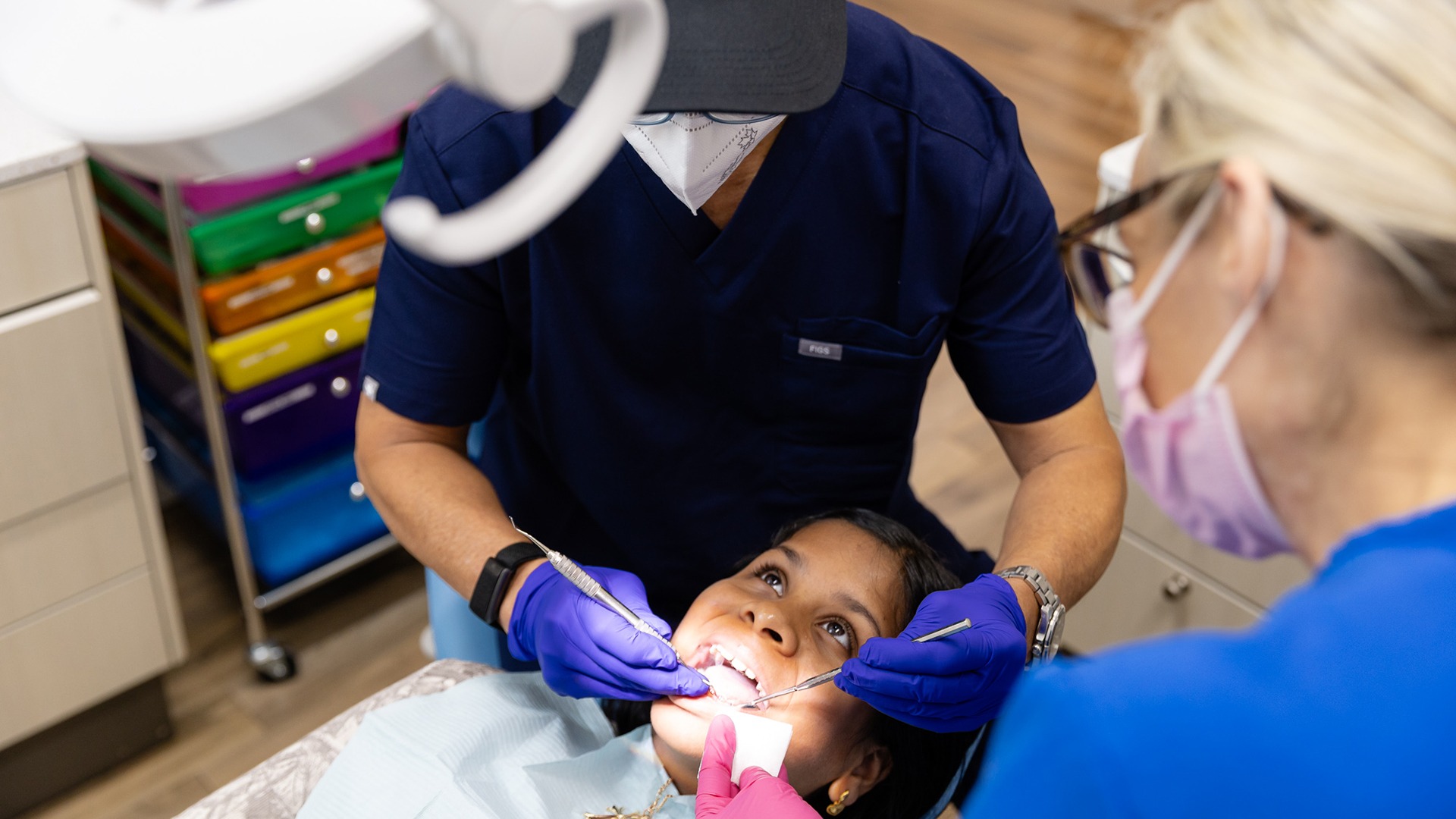 A child receives dental care from two persons wearing gloves and masks. Colorful storage units are visible in the dental office setting.