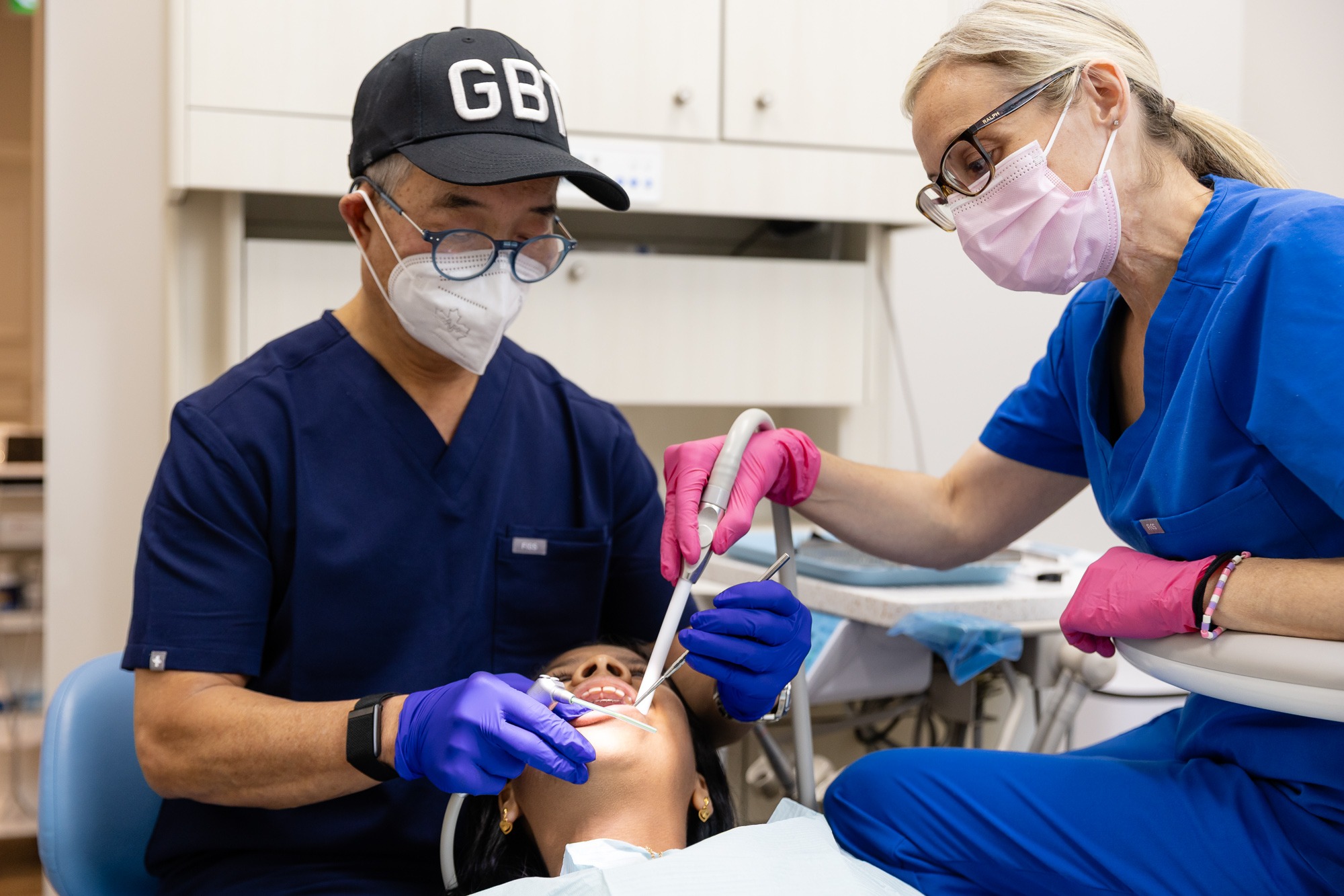 Two persons in medical attire perform a dental procedure on a patient lying in a dental chair. They wear gloves and face masks.