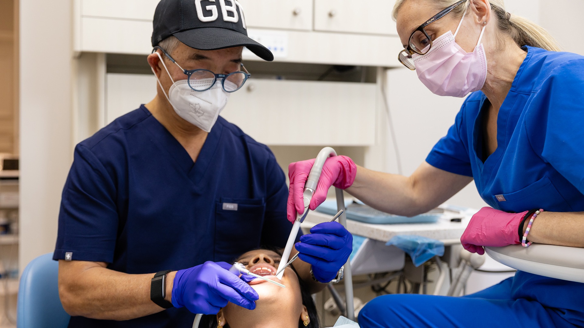 Two dental professionals wearing masks and gloves conduct a procedure on a patient in a clinic setting, focusing on oral hygiene.