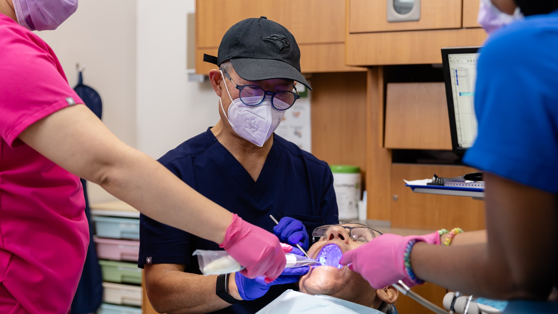 Three dental professionals attend to a person in a dental clinic, using various instruments. The setting is clean, organized, and well-equipped.