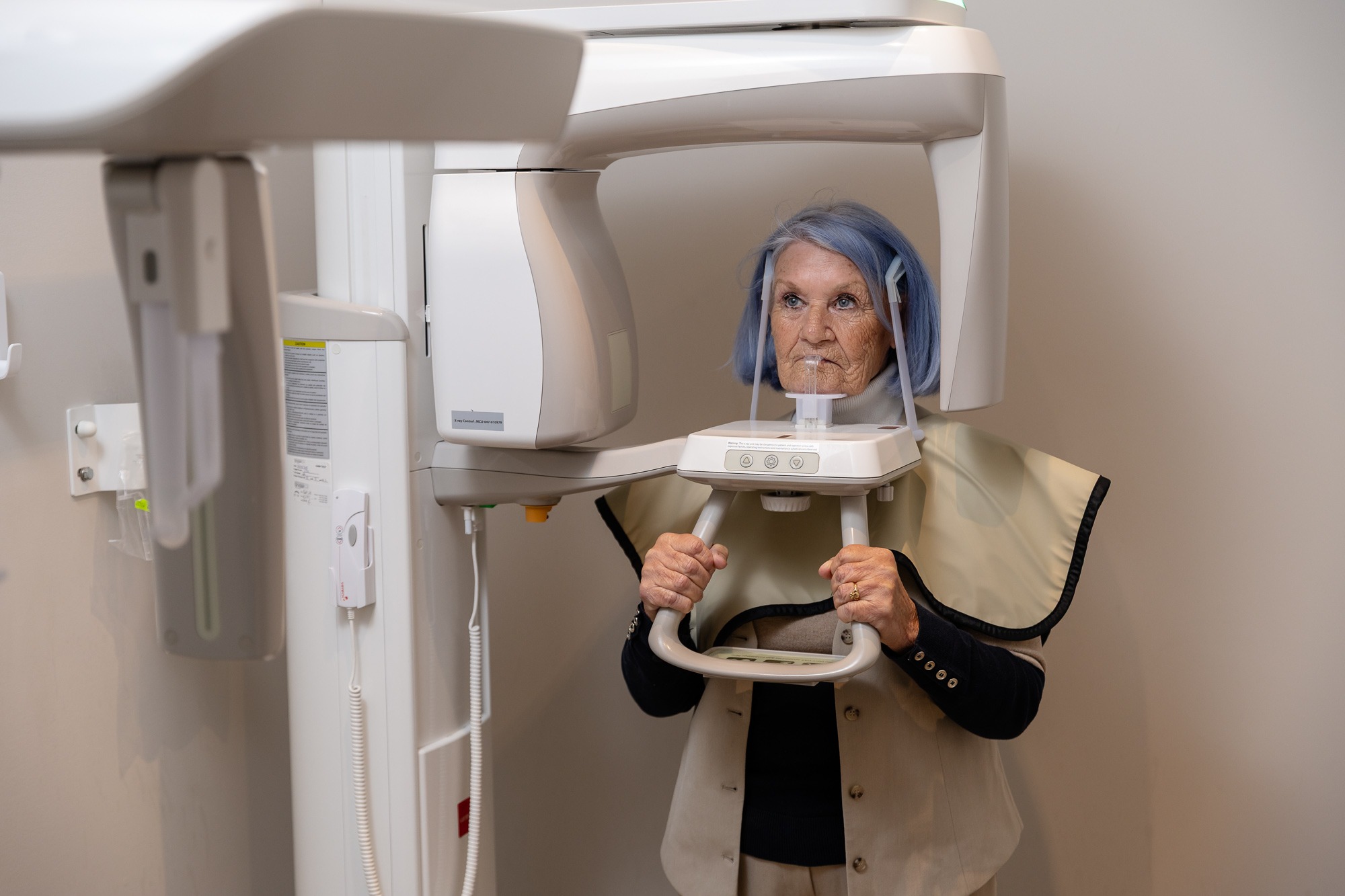 A person with blue hair undergoes a dental X-ray scan, standing in medical equipment, wearing a protective vest in a plain room.