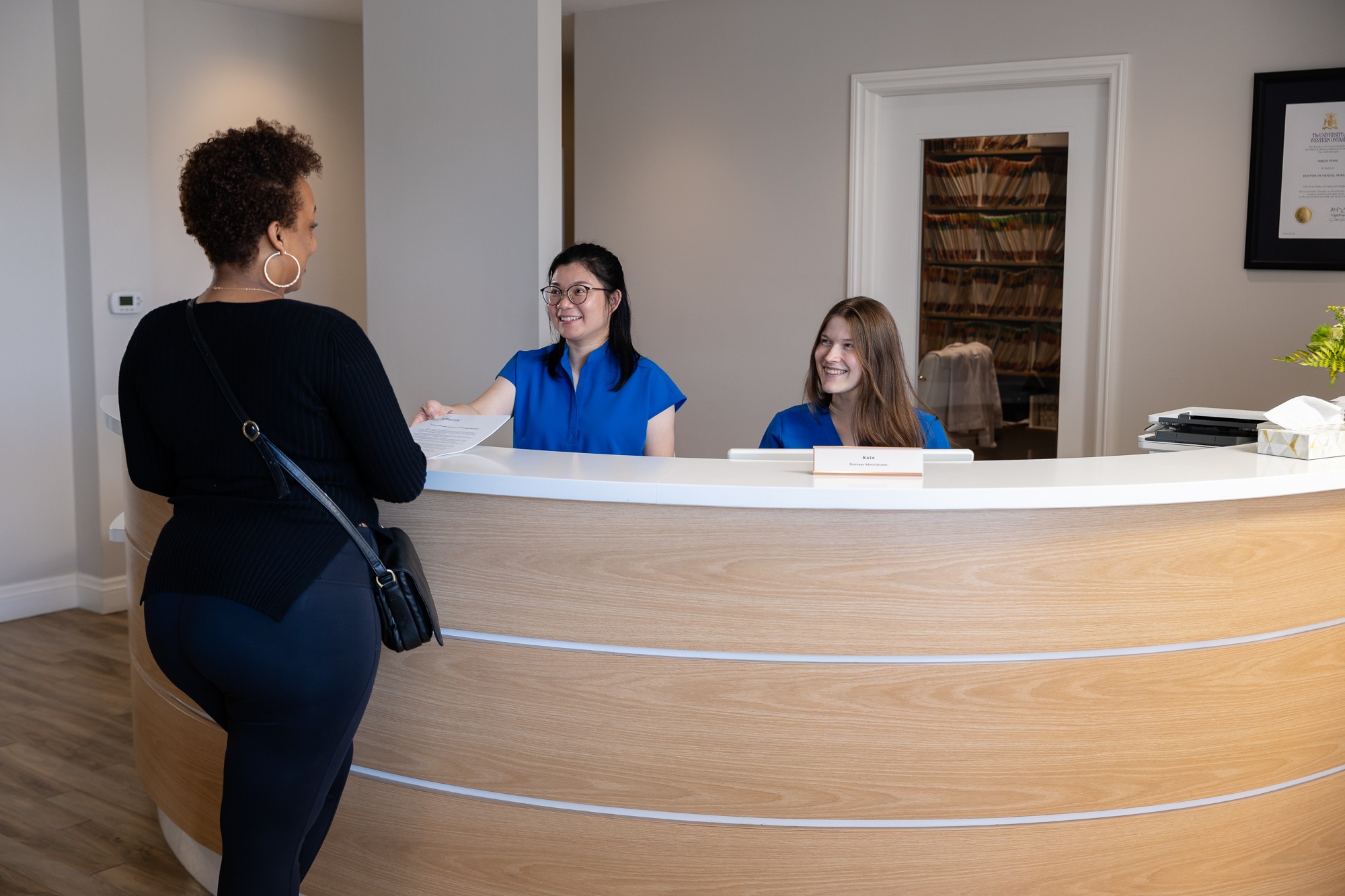 A person at a reception desk interacts with two staff members. The environment is professional and welcoming, featuring office decor and paperwork.