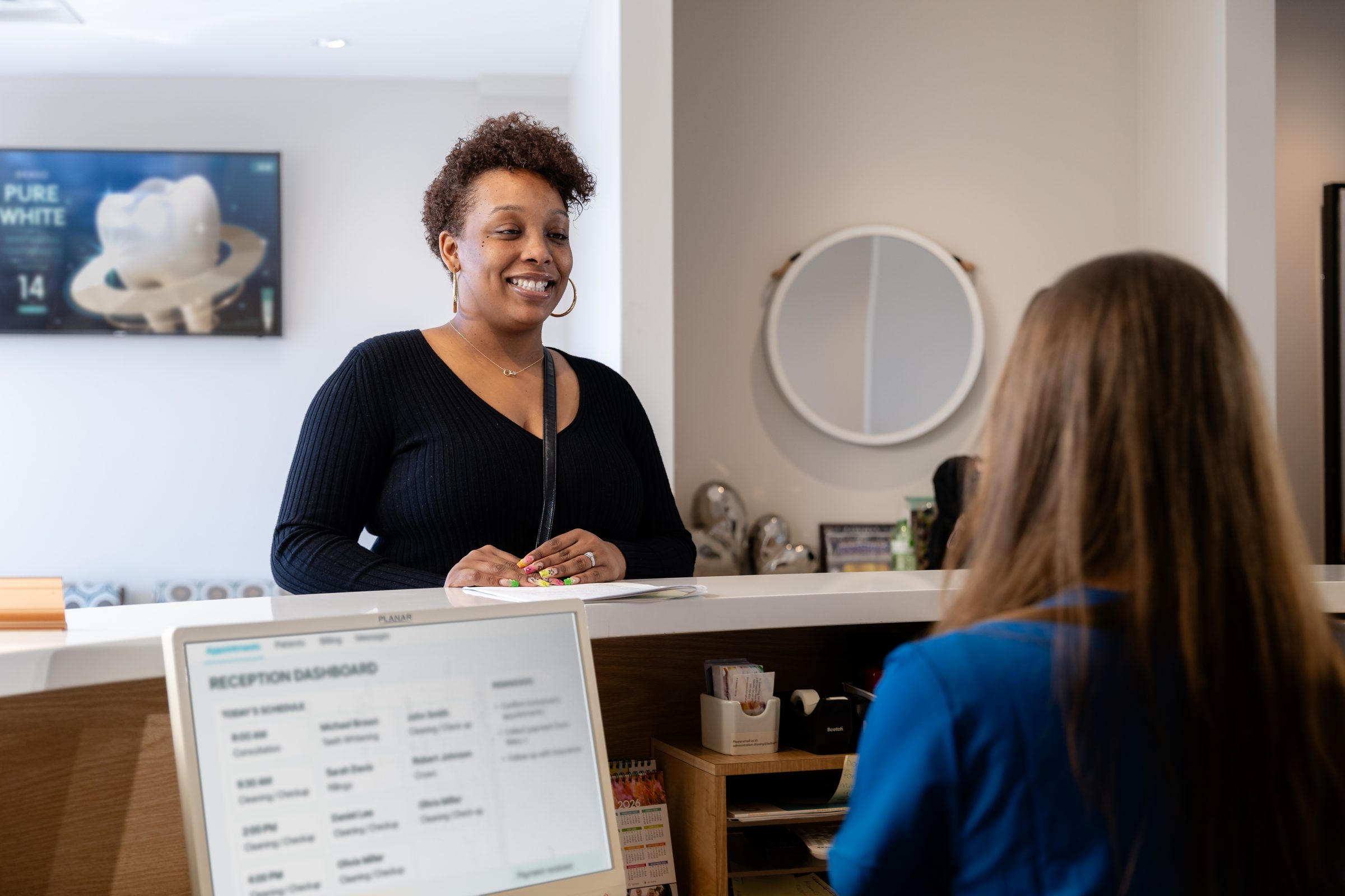 A person smiles at a reception desk, engaging with another person. A computer screen and promotional poster are visible in the background.
