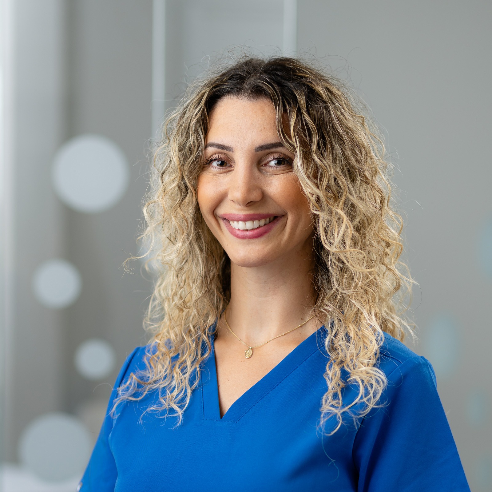 A person with curly hair smiles, wearing a blue top, standing in a softly lit, blurred background with circular light reflections.