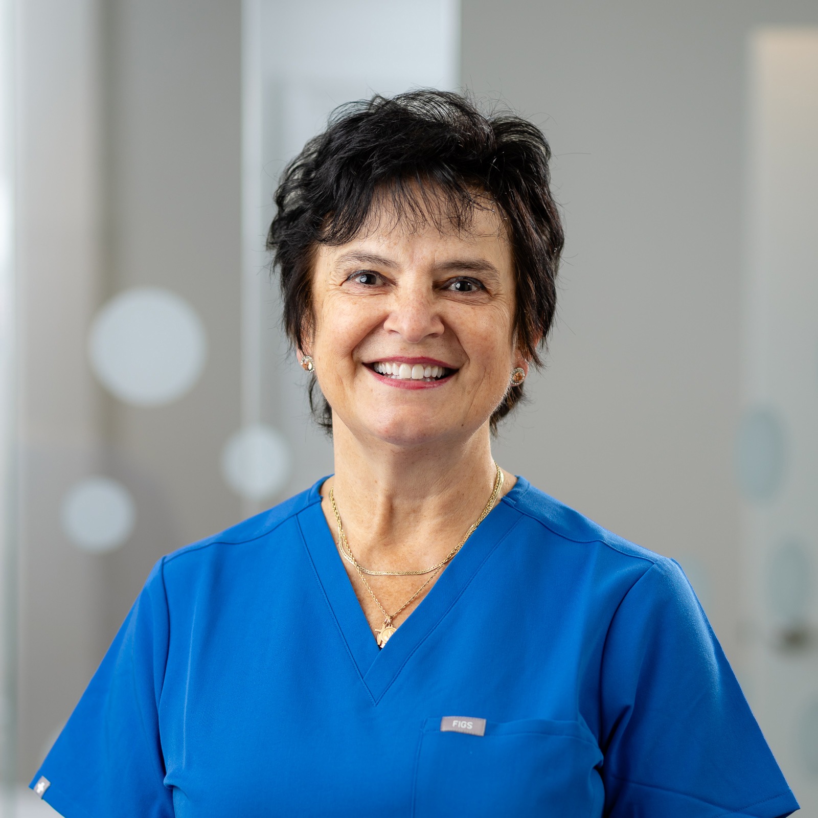 A smiling person wearing blue scrubs stands indoors with a blurred modern background, featuring circular light reflections on glass.