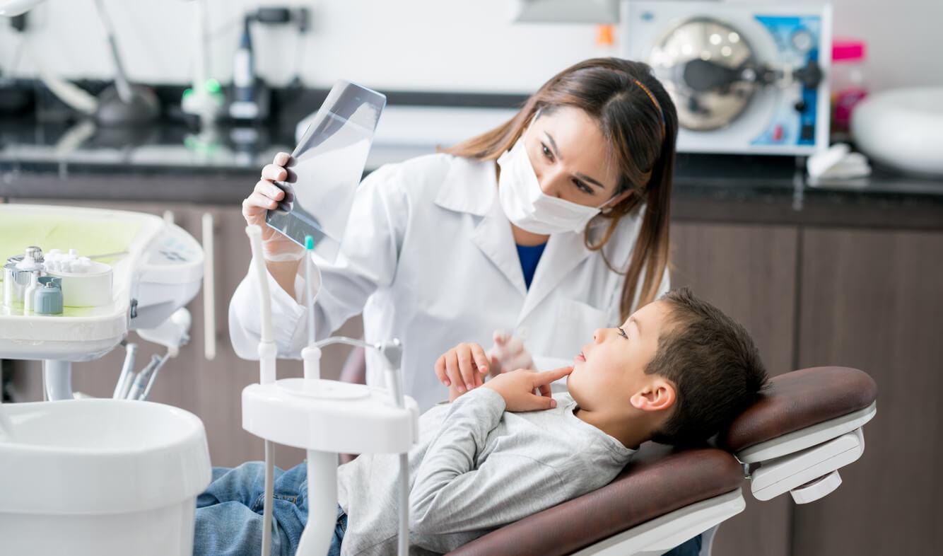 A person in a dental office shows an X-ray to a child sitting in a dental chair. Equipment is visible around them.