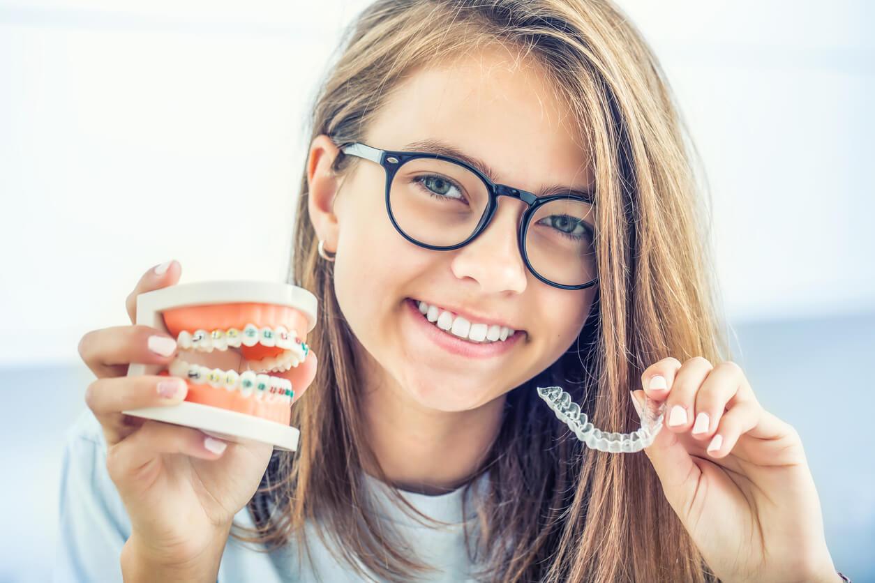 A person wearing glasses smiles, holding a dental model with braces and a clear aligner, demonstrating orthodontic appliances in a bright setting.
