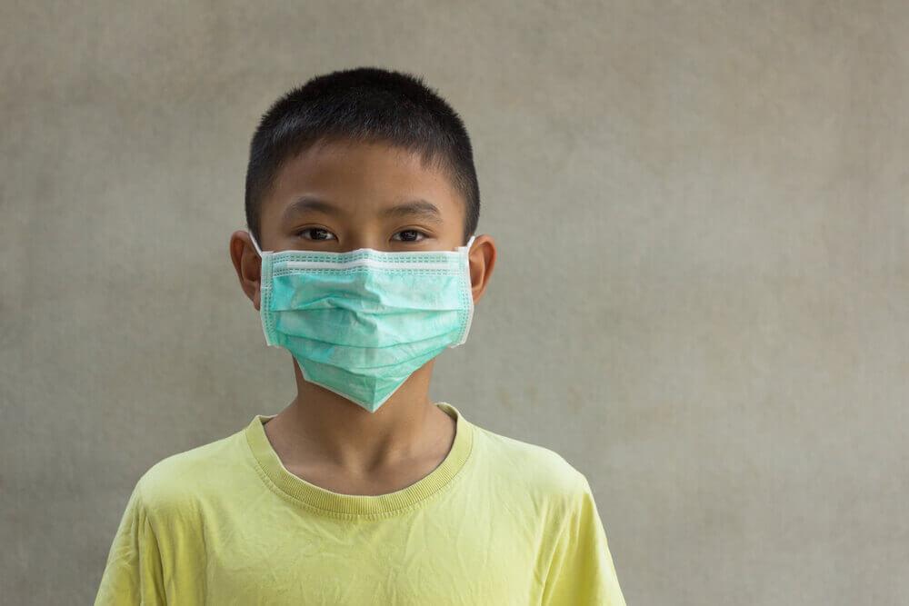 A child wearing a medical mask stands against a plain background. The child wears a light yellow shirt and looks directly forward.
