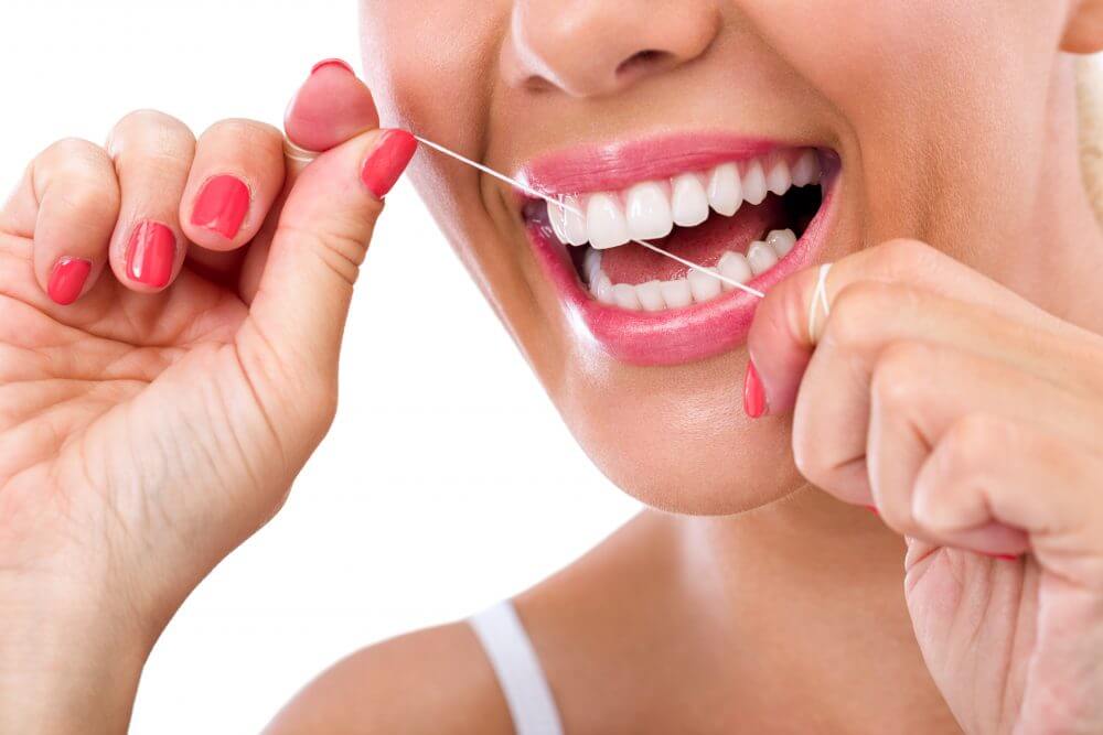 A person with red nails is smiling while flossing their teeth, displaying healthy white teeth and vibrant pink lips.