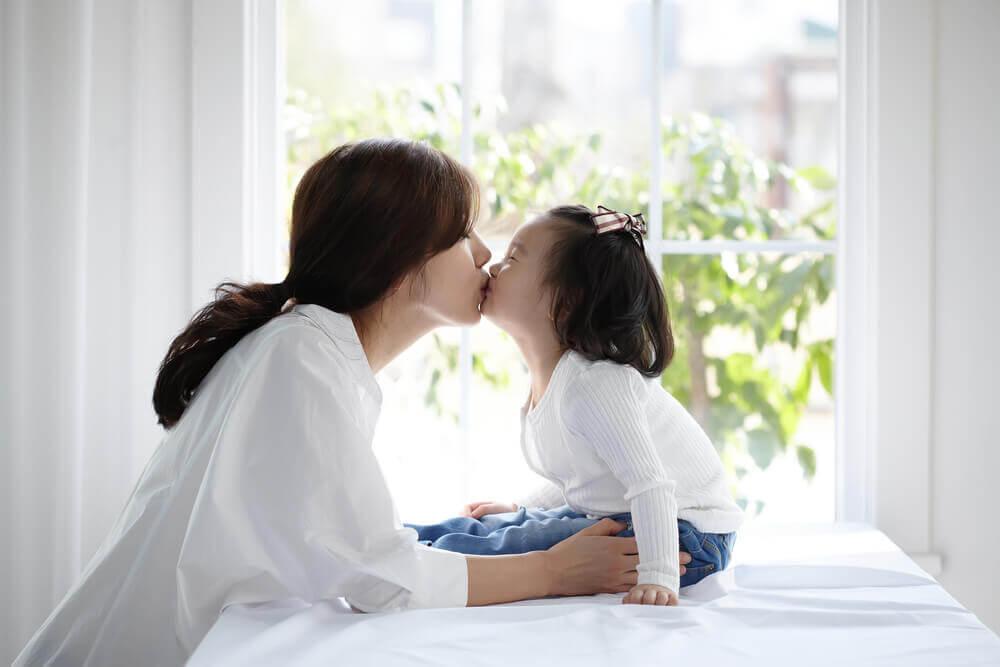 A person and child share a tender moment, kissing by a sunlit window with greenery visible in the background.