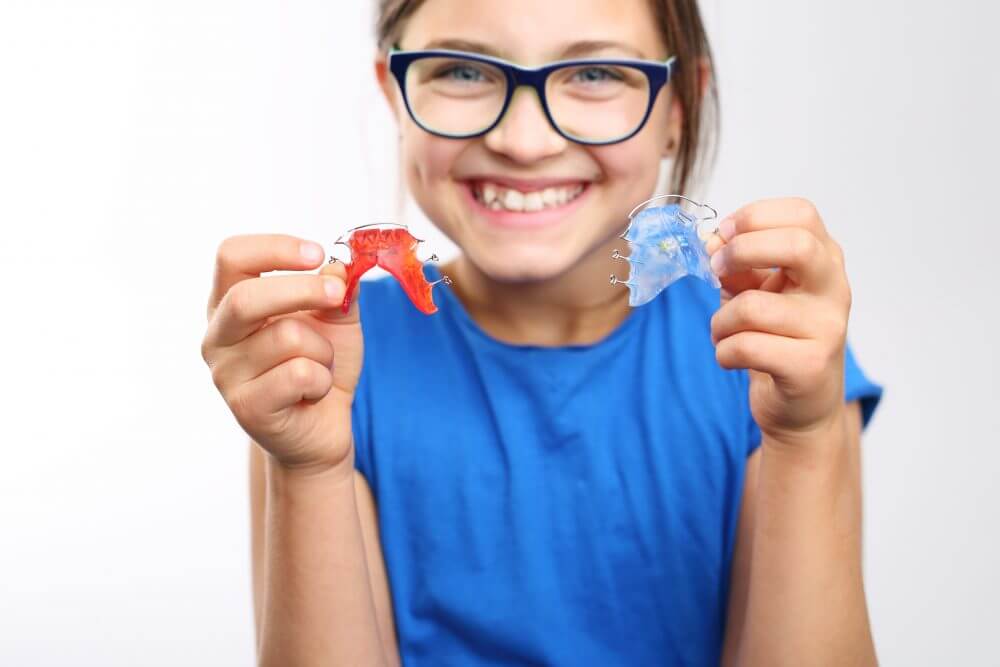 A smiling child holds colorful dental retainers, one red and one blue, against a plain background. The child wears blue and glasses.