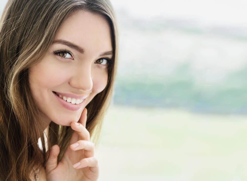 A person with long hair smiles warmly, touching their chin gently, set against a softly blurred, serene outdoor background.