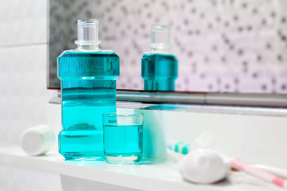 Bottle of blue mouthwash and glass sit on a bathroom counter, reflected in the mirror. Nearby are toothpaste and a toothbrush.