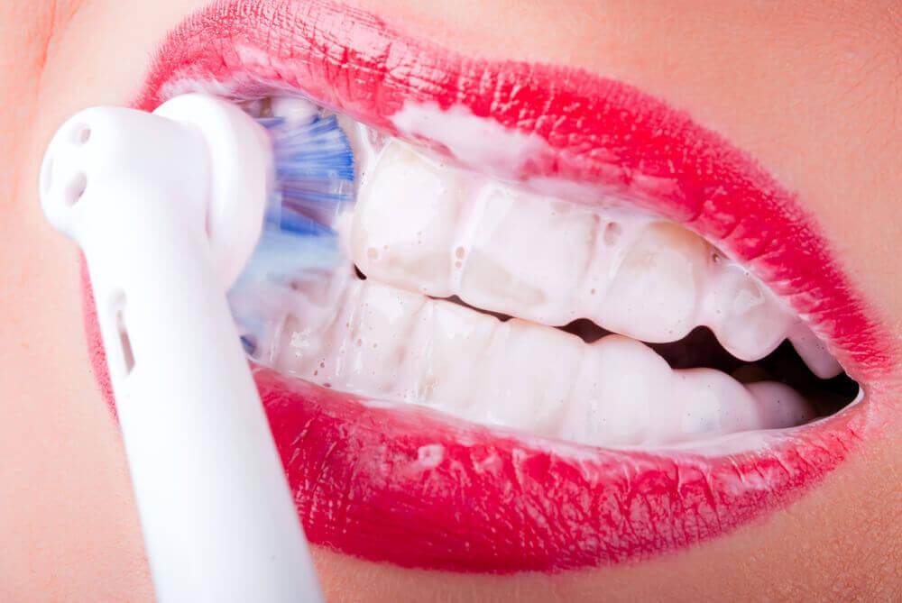 A close-up of a person’s mouth with red lips, showing teeth being brushed with an electric toothbrush and white toothpaste foam.