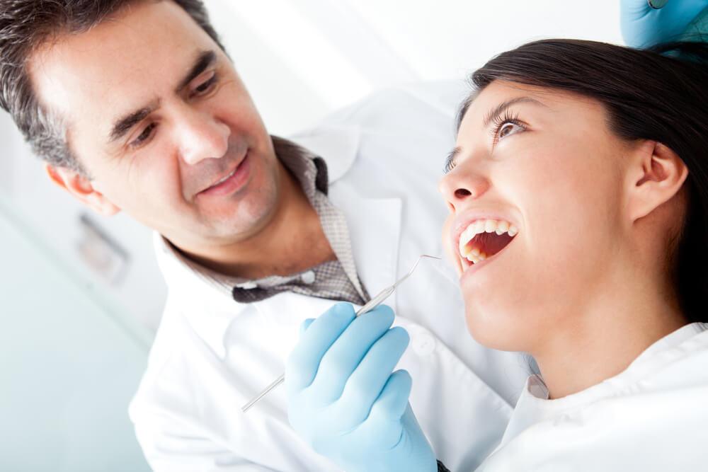 Two people are in a dental office. One person examines the other's teeth with dental tools while both smile.
