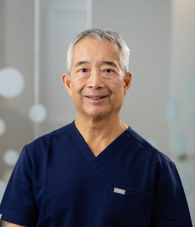 A person wearing navy blue scrubs stands indoors, smiling against a blurred background featuring abstract circular patterns.