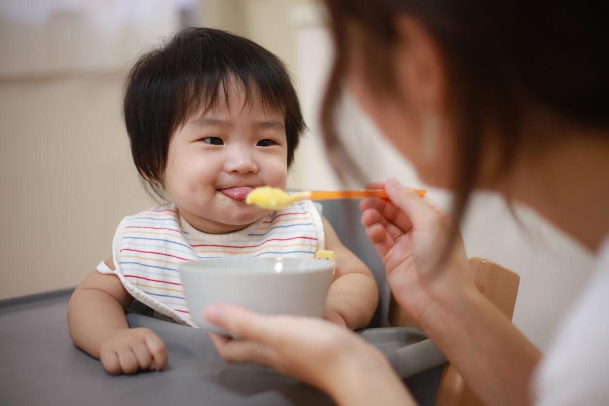 A person feeds a smiling baby in a high chair, using a spoon. The baby wears a striped bib and shows playful expression.