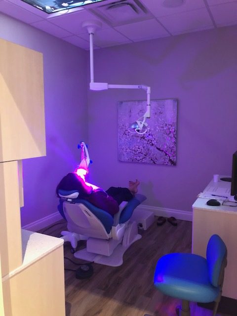 A person is seated in a modern dental office, receiving treatment under purple light, surrounded by pastel walls and a floral painting.