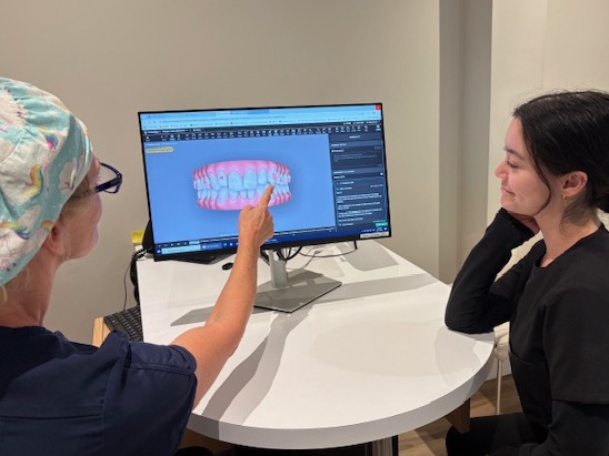 Two people in a dental office, one explaining digital teeth model on a screen to the other. They are seated at a round table.