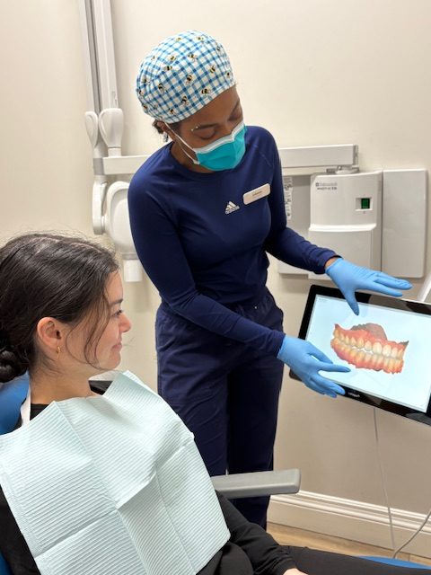 Two people in dental office; one seated in chair, other showing teeth scan on monitor, both wearing masks and gloves.