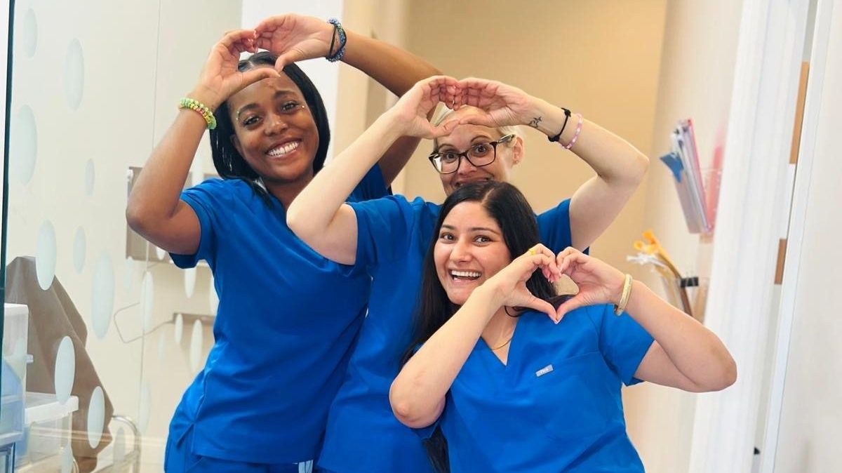 Three people in blue scrubs make heart shapes with hands, smiling in a clinical setting. White walls and medical folders visible.